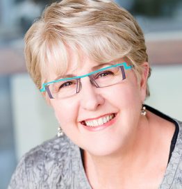 Bio portrait of Brenda Collier, smiling in a silver shirt, with turquoise-rimmed glasses